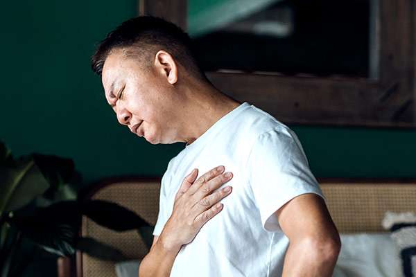 Close up of senior Asian man holding his chest in discomfort, suffering from chest pain while sitting on bed at home.